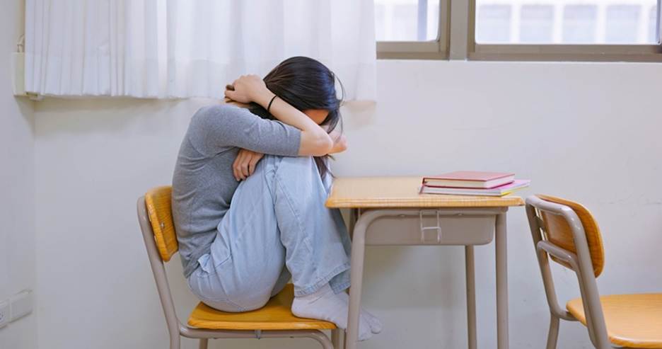 A despondent student sits hunched in a school desk with head in arms. The scene is likely about issues of sexual abuse on campus.